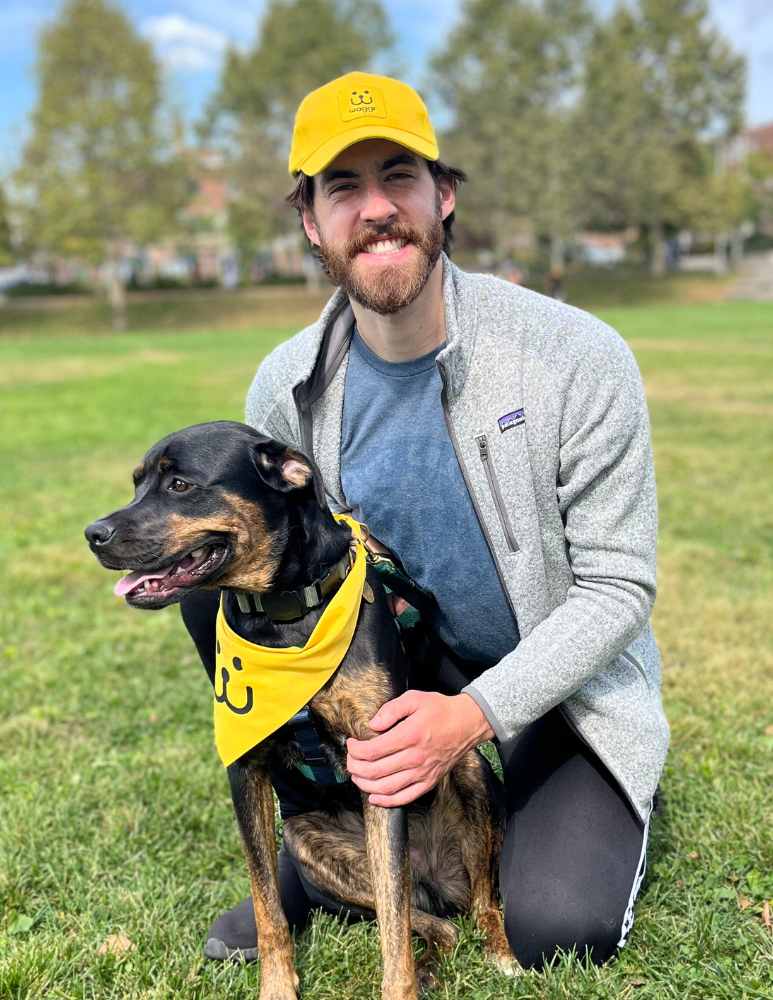 Smiling man wearing yellow waggr cap holds a rottweiler wearing a yellow bandana.